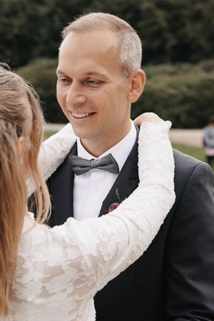 Smiling groom in black tuxedo and gray bow tie with boutonniere as bride in lace dress adjusts his lapel outdoors.