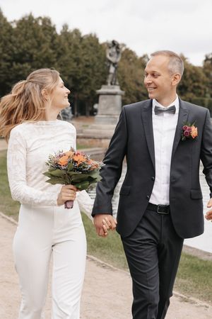 Happy bride and groom walking hand in hand outdoors, bride holding bouquet, groom in black tuxedo with boutonniere.