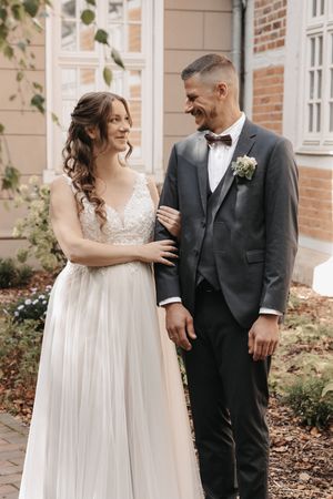 Bride in white lace gown and groom in gray suit with bow tie share a joyful moment outside a brick building.