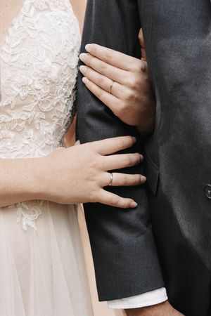 Bride in lace gown rests both hands on groom's gray suit sleeve, showing off wedding rings.