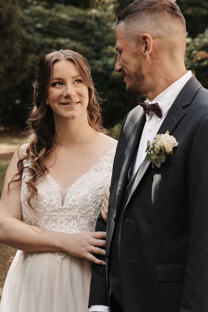 Bride in lace wedding gown smiles at groom in dark suit with boutonniere outdoors.
