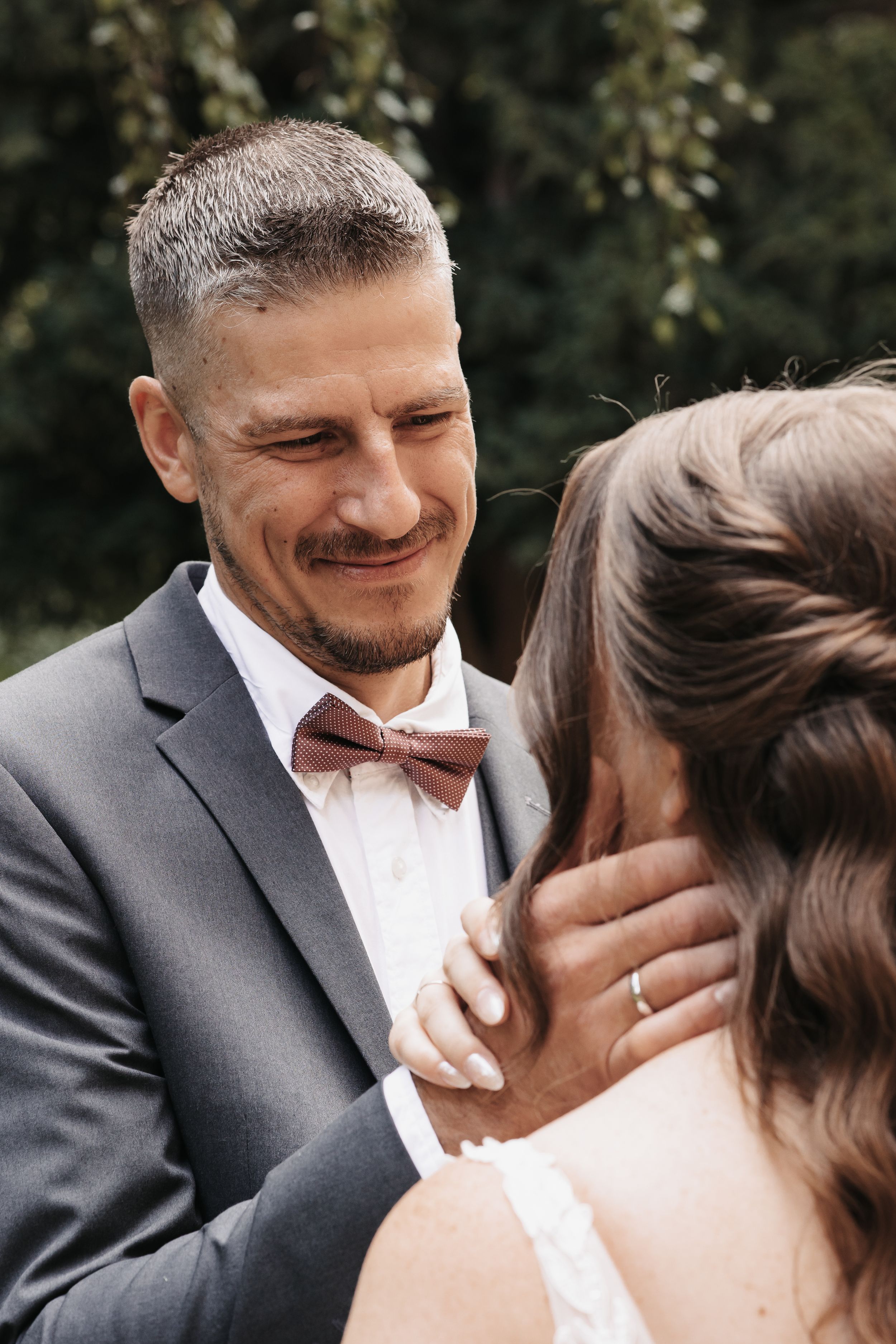 Groom in gray suit and mauve bow tie smiling tenderly at bride during outdoor wedding ceremony.