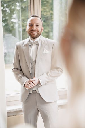 Smiling groom in a light beige suit with bow tie standing by a bright window on his wedding day.
