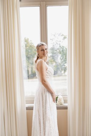 Smiling bride in white lace gown standing by large window with cream curtains, holding white flowers.