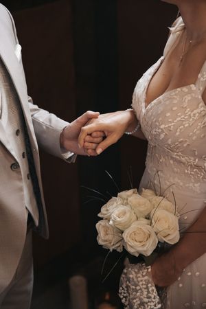 Bride and groom holding hands at wedding ceremony, bride holding white rose bouquet, wearing lace gown and gray suit.