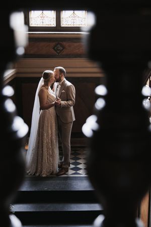 Bride and groom share an intimate moment on ornate staircase, framed by stained glass windows in elegant venue.
