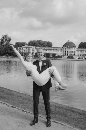 Groom carries laughing bride in wedding attire by a lake with a grand building backdrop in black and white.