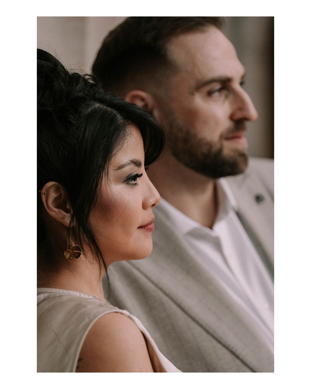 A bride and groom in profile, woman with dark updo and elegant makeup, man with beard in gray suit.