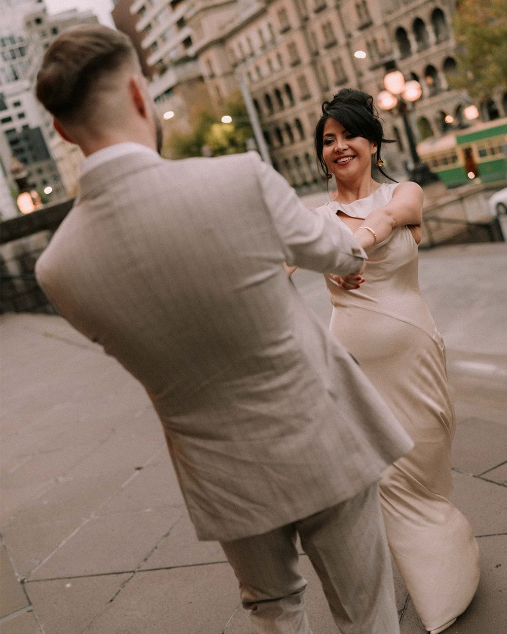 A bride in a satin gown dances with a groom in a beige suit on a city street during their wedding.