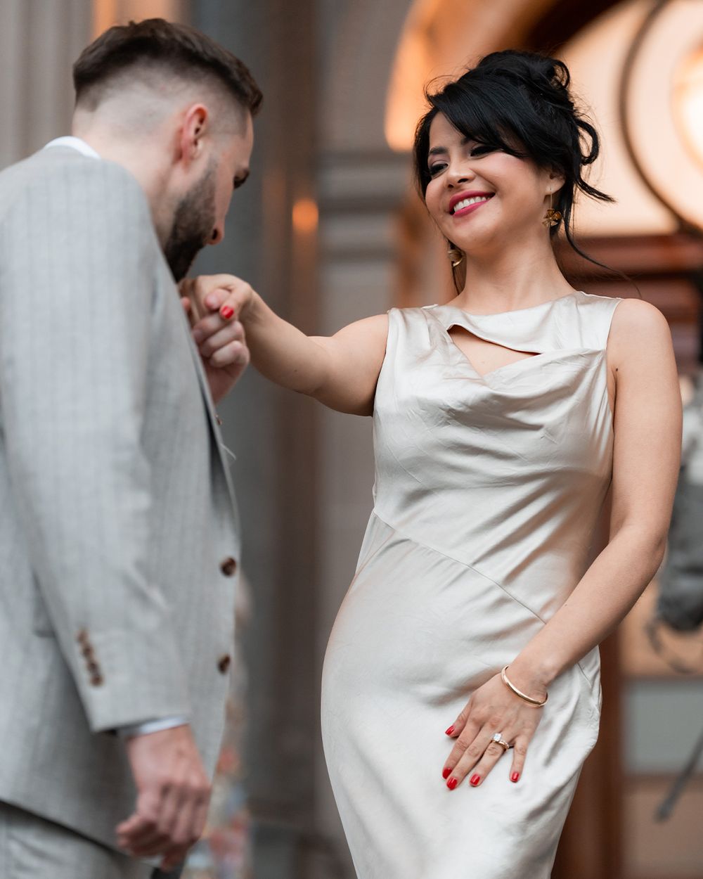 A man in a gray suit kisses a woman's hand as she smiles, wearing a white dress with red nails.