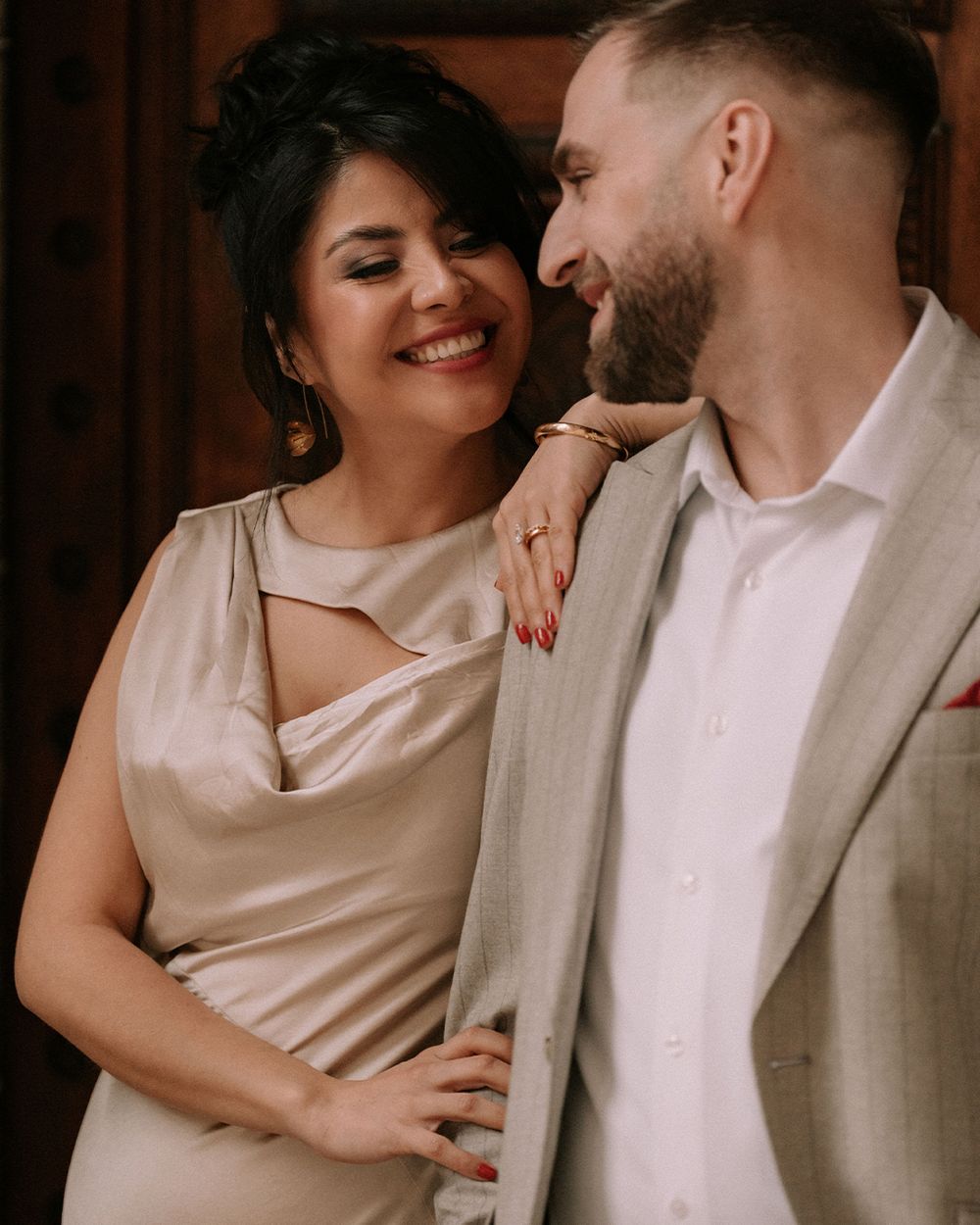 A smiling woman in a beige satin dress leans against a man in a linen blazer, sharing a romantic moment.