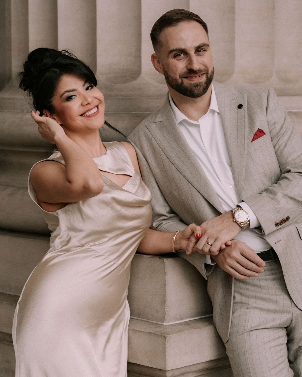 Smiling couple in elegant wedding attire posing together against stone columns.