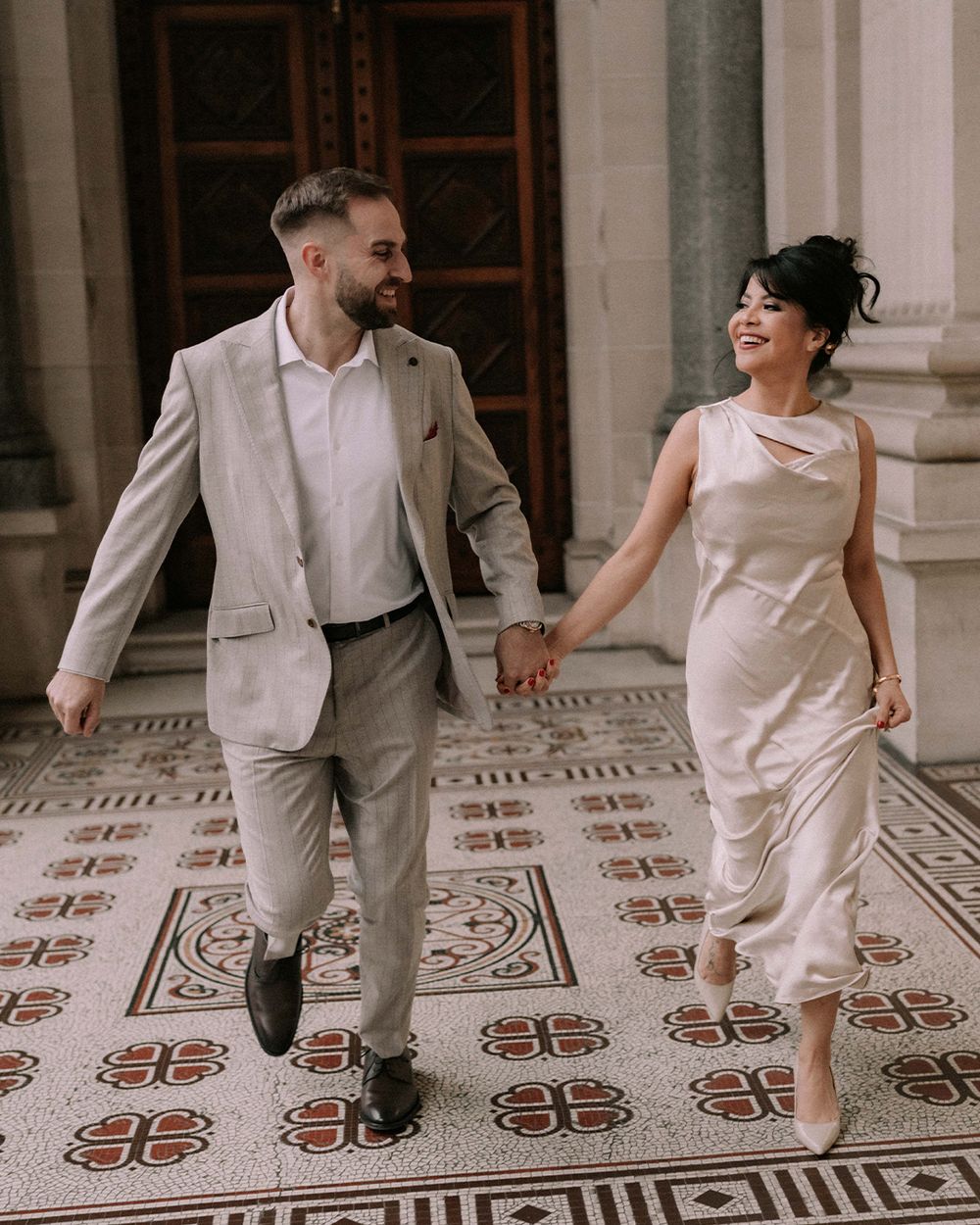 Couple holding hands and walking together in elegant wedding attire on a decorative tiled floor.