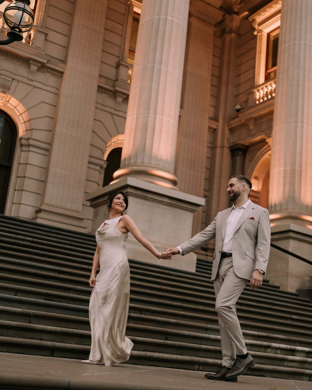Couple holding hands on grand stone steps of a classical building, bride in white gown, groom in beige suit.