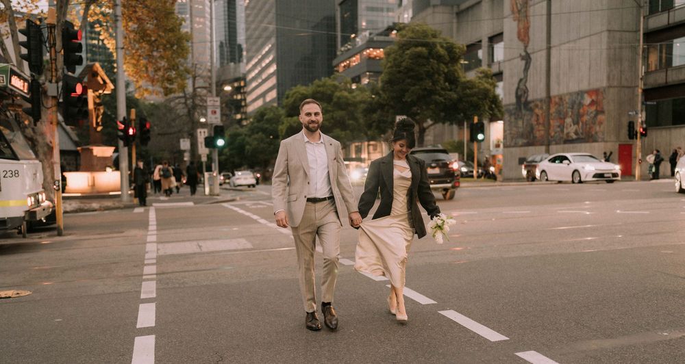 Couple holding hands crossing a city street, man in beige suit and woman in white dress, urban skyline backdrop.