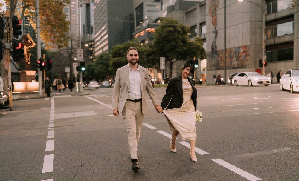 Couple holding hands crossing a city street, man in beige suit and woman in white dress, urban buildings in background.
