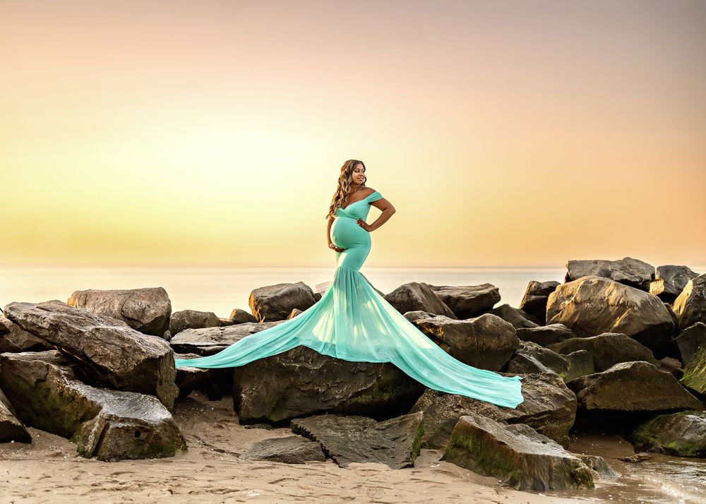 A turquoise evening gown flows elegantly across dark coastal rocks at sunset creating a dramatic fashion scene.