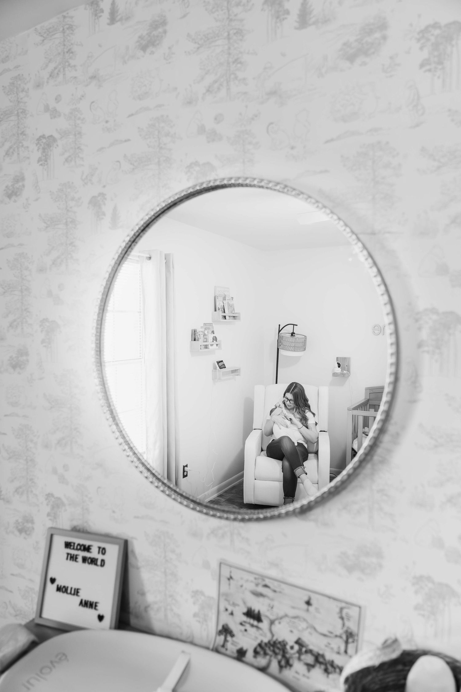 A black and white photo of a circular mirror on a textured wall, with artwork and decorations visible below it.
