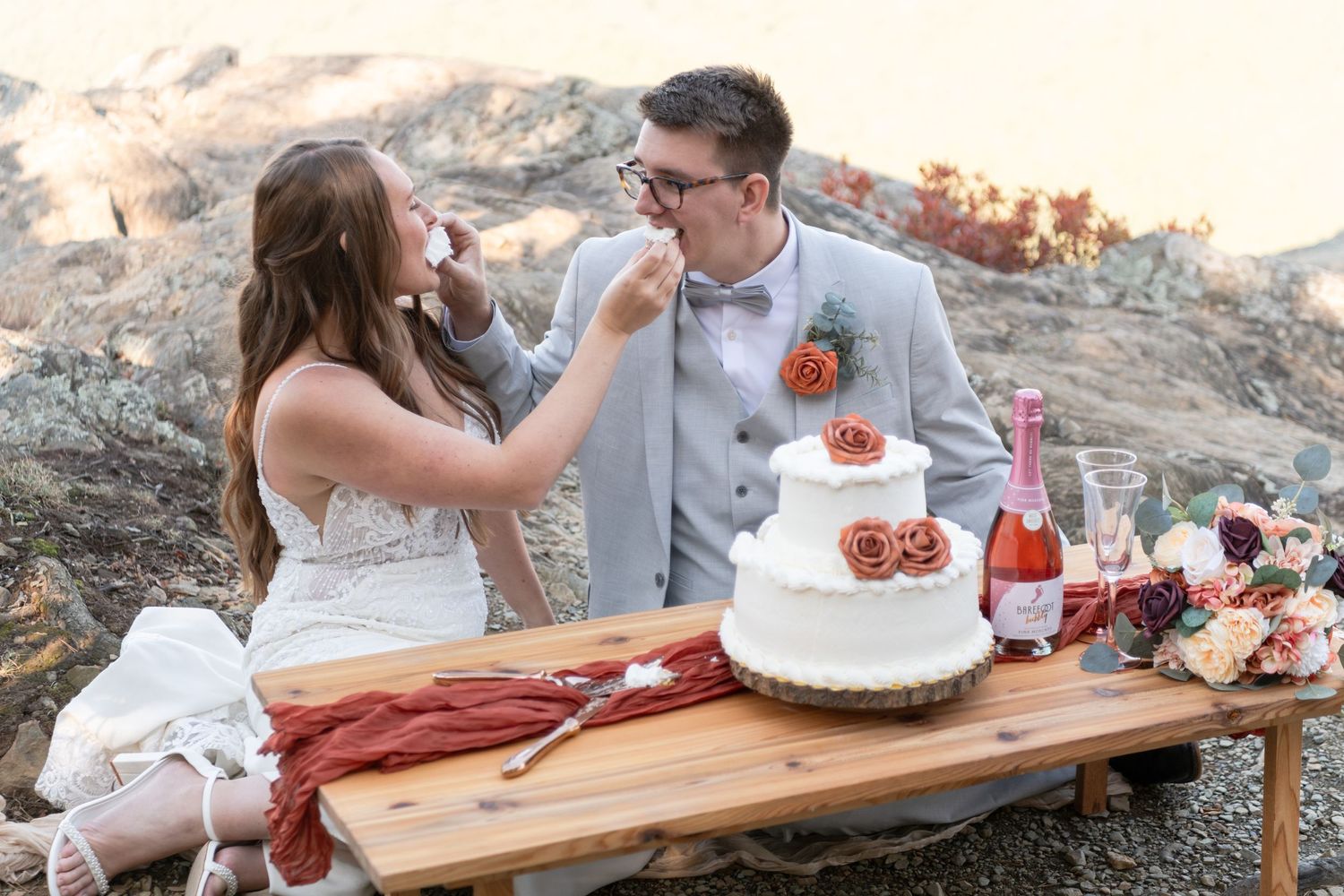 A couple shares wedding cake during a sunset picnic on a wooden board with champagne and strawberries.