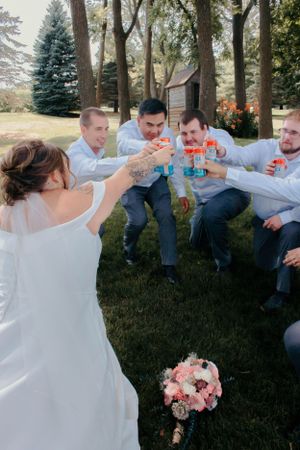 Wedding party members in white shirts toast with colorful drinks outdoors.