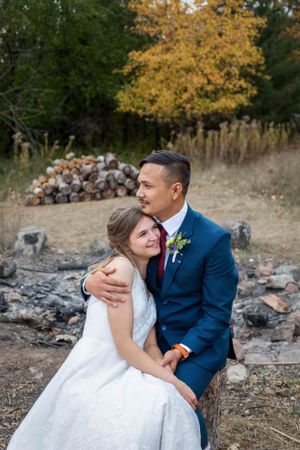 A couple in wedding attire embrace on rocky terrain with fall foliage in the background.