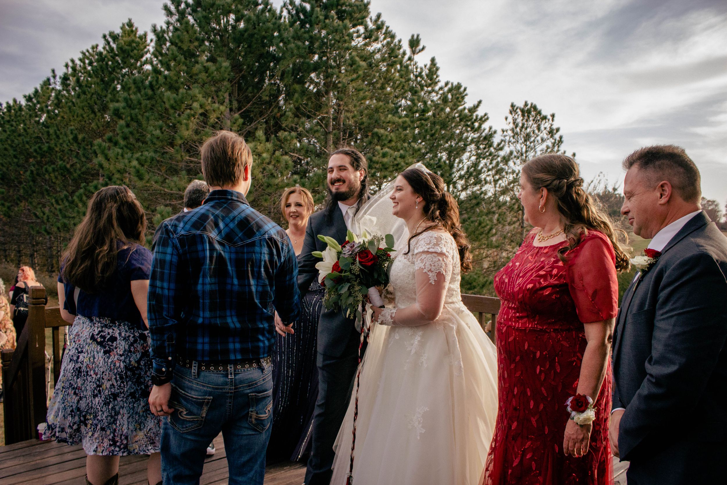 A group gathers outdoors for a wedding ceremony with evergreen trees in the background and cloudy skies above.