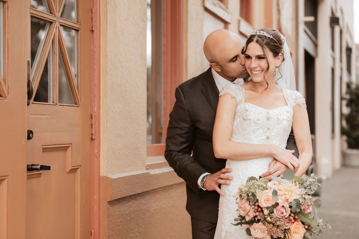 A tender wedding moment captured outside a building with coral-colored doors.