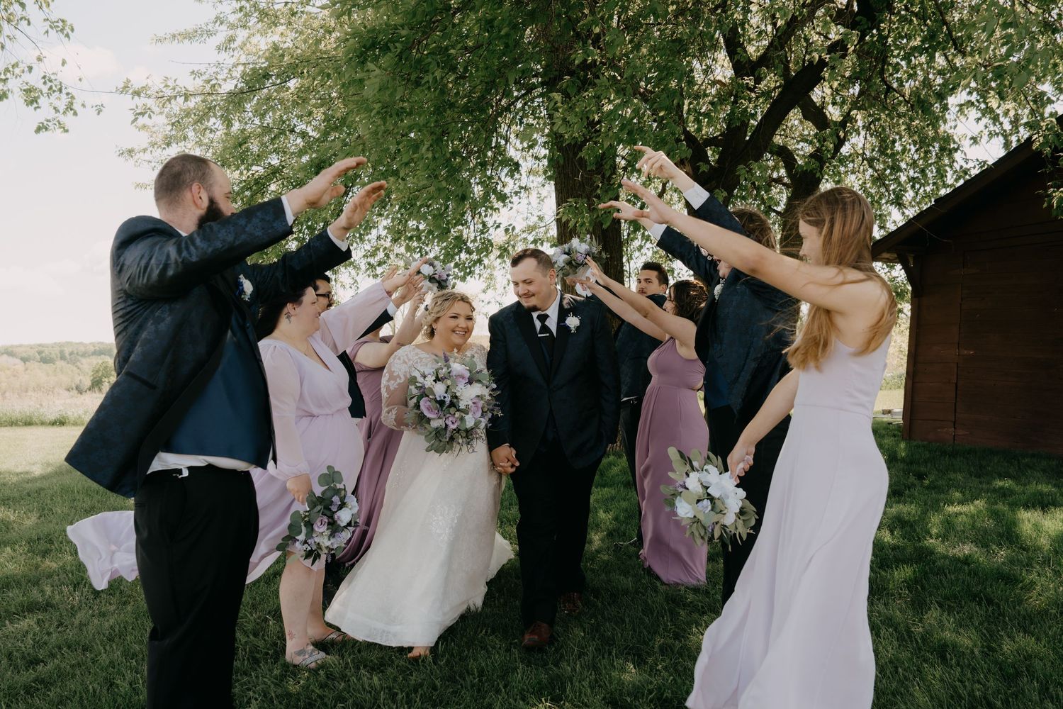 Wedding party celebrates outdoors with champagne toasts near a rustic barn on a sunny day.