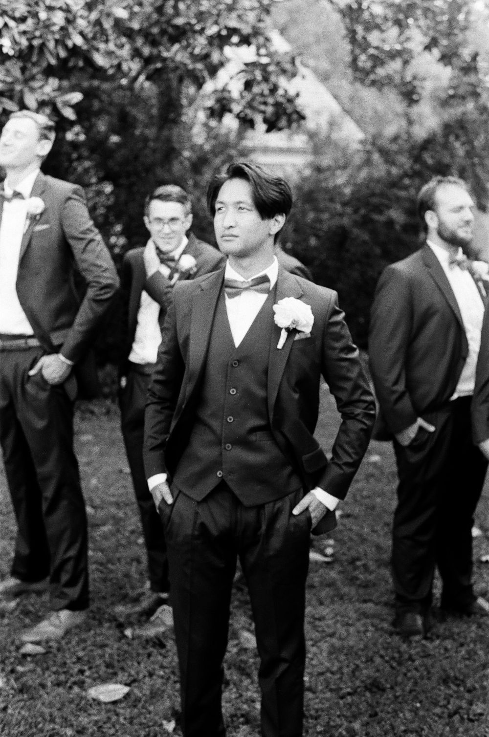Black and white portrait of groom in three-piece suit with boutonniere and groomsmen behind.