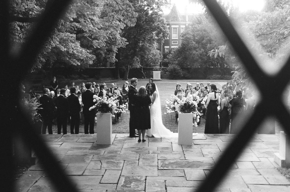 Outdoor wedding ceremony viewed through decorative iron gate with guests and floral arrangements.