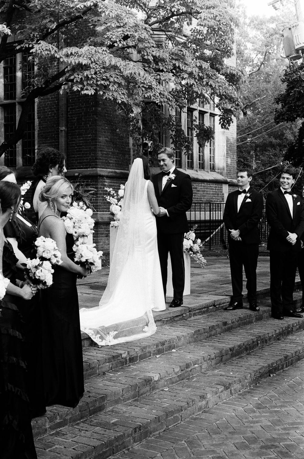 Black and white photo of bride approaching groom at altar on brick steps with wedding party.