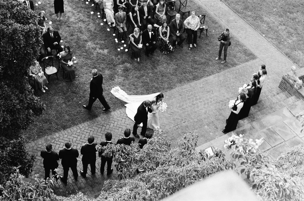 Aerial view of wedding procession with bride and groom walking on brick pathway.