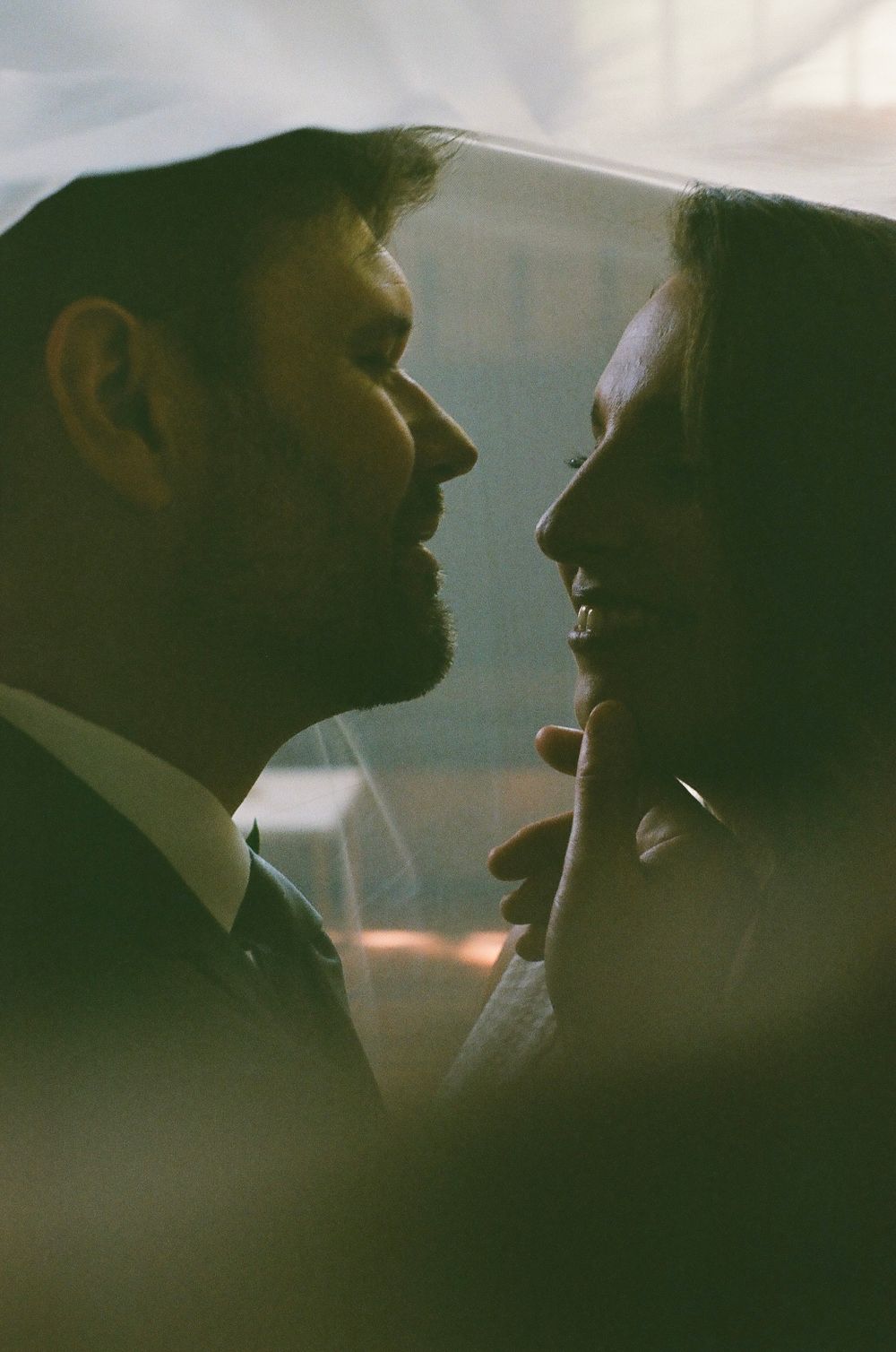 Romantic silhouette of bride and groom facing each other under a veil in soft light.