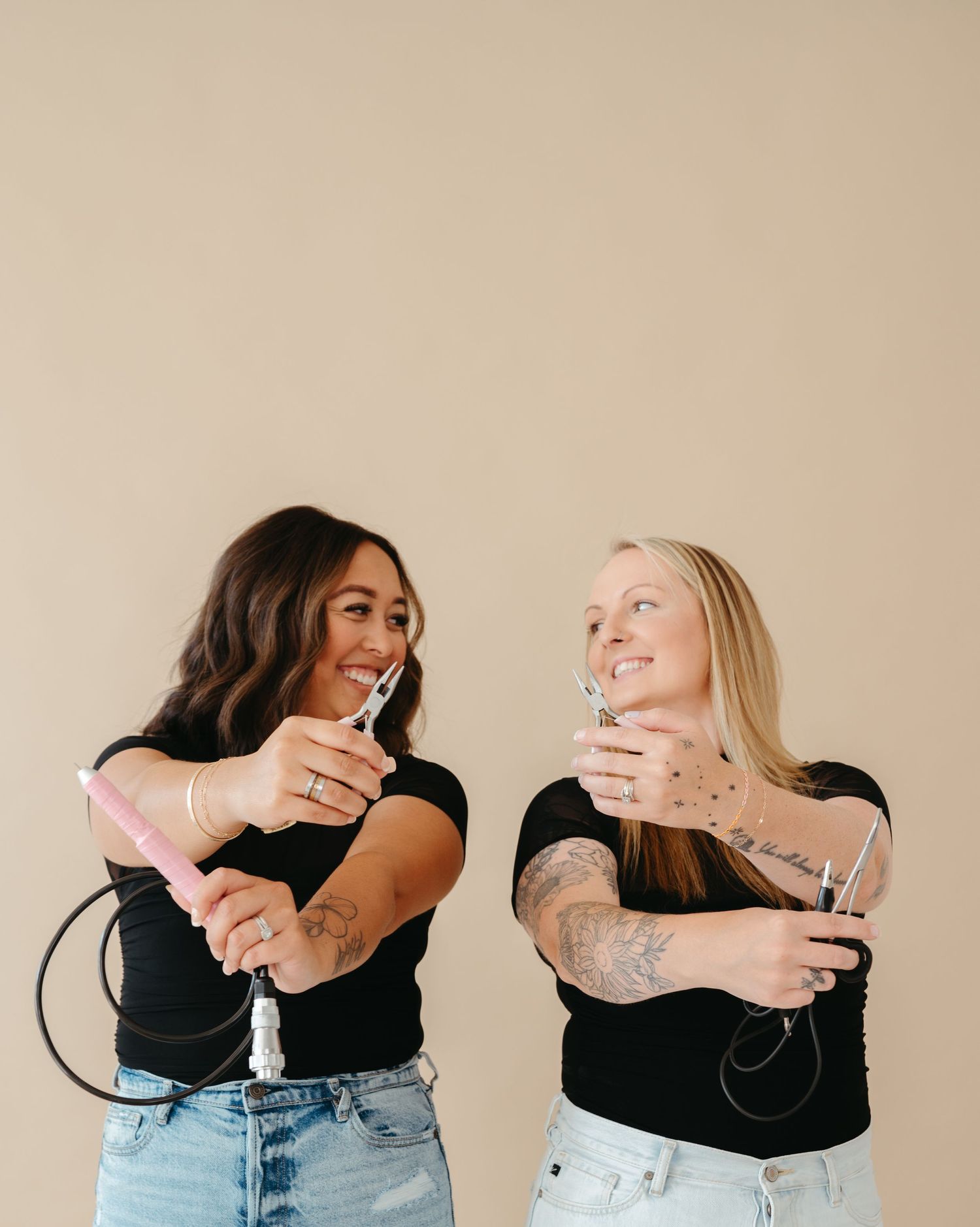 Two friends in black shirts and jeans share a joyful moment together against a beige backdrop.