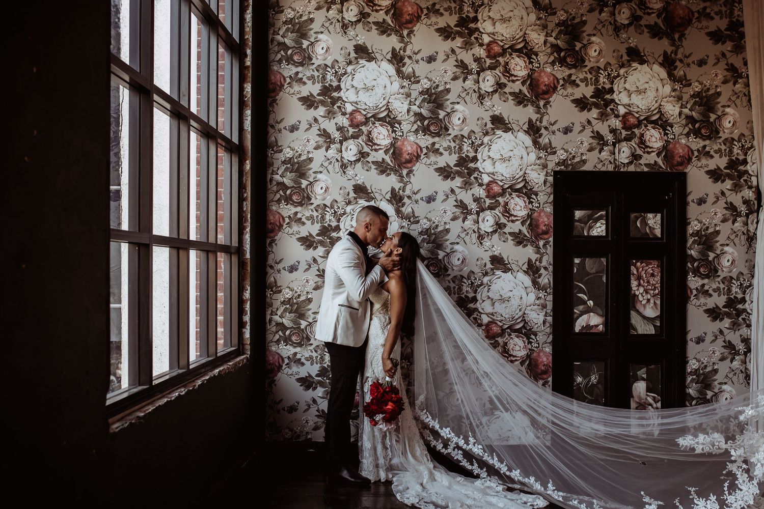A bride in a white gown with a long veil stands by a window against a vintage floral wallpapered wall.