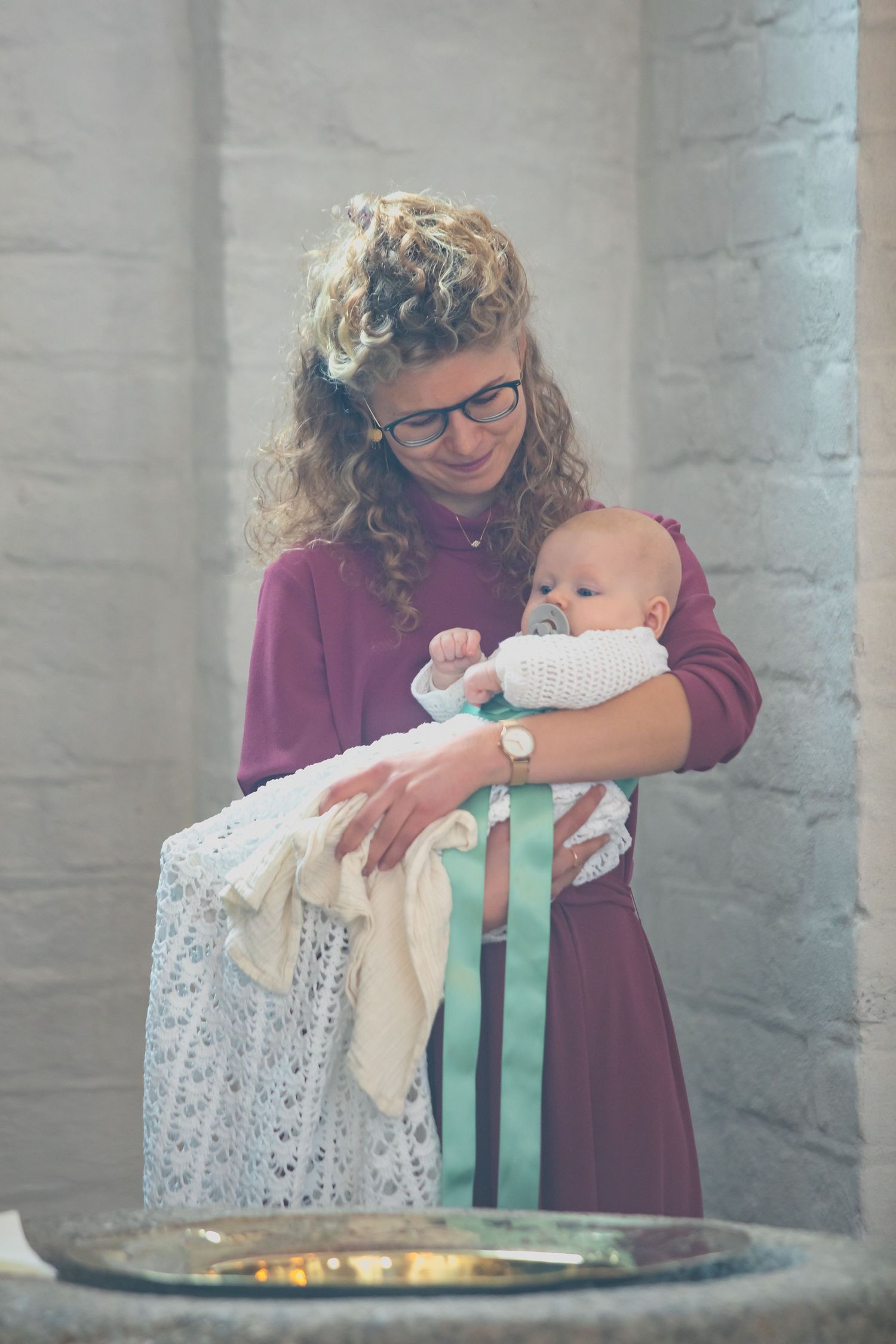 A christening ceremony showing a burgundy-dressed person holding an infant in white lace clothing.