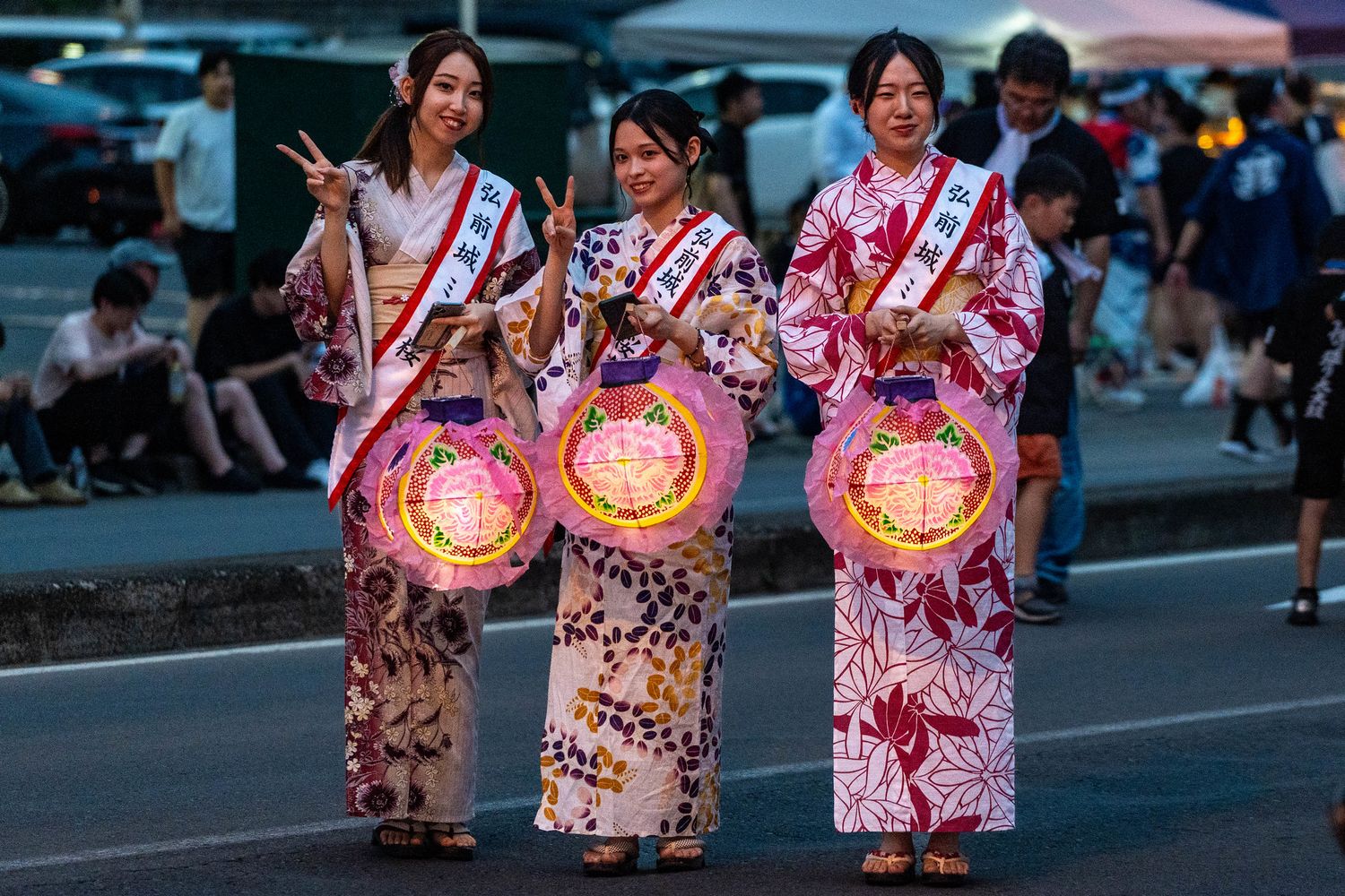 Three participants in colorful yukata holding glowing lanterns at a summer festival.