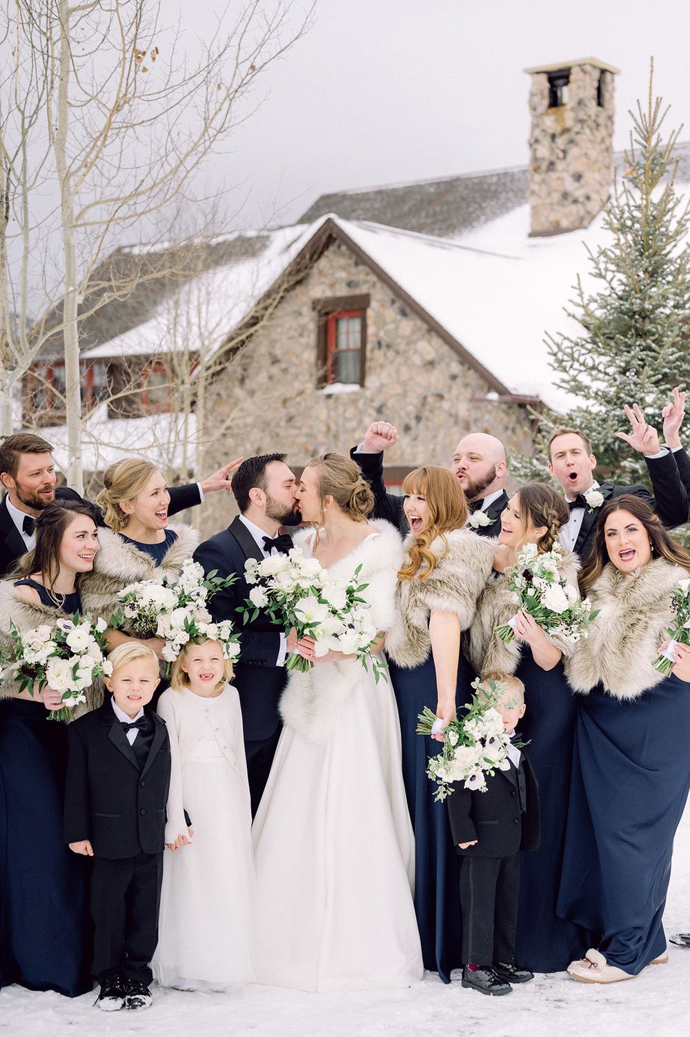 A wedding party poses together in front of a snow-covered stone cottage during winter.
