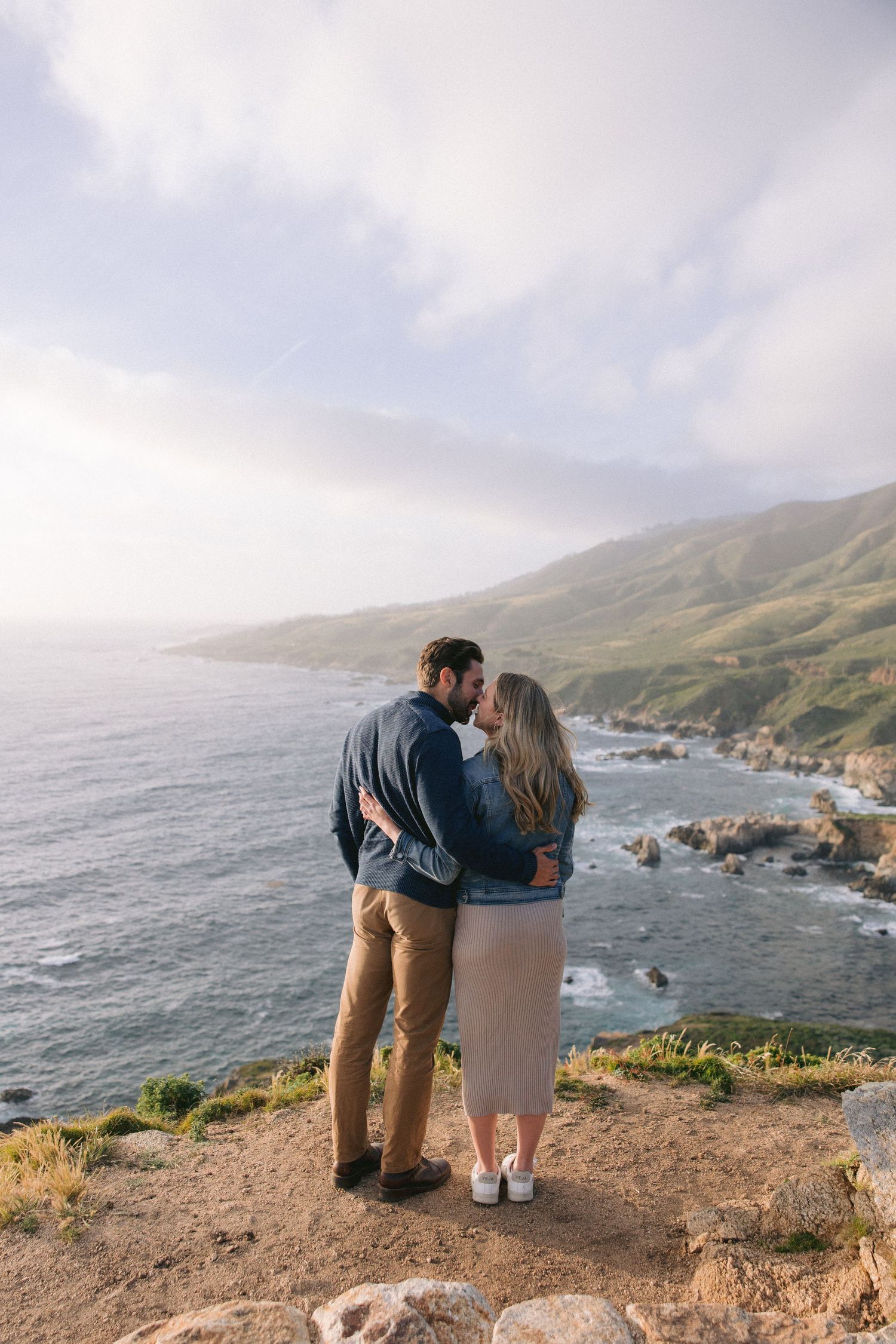 A couple embraces while admiring the dramatic coastal views along Big Sur's rugged cliffs at sunset.