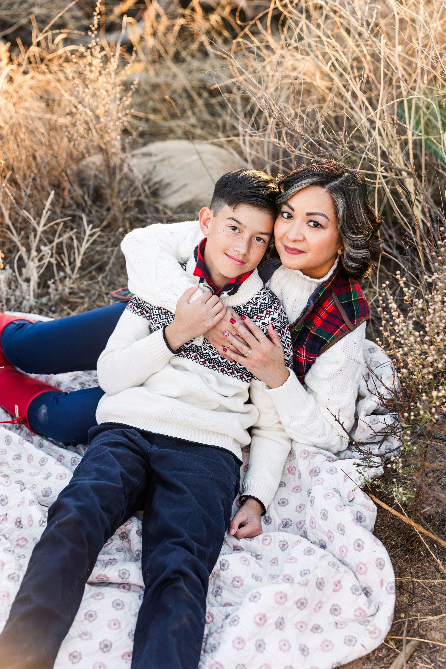 A couple in cozy winter sweaters relaxes on a patterned blanket outdoors among dry autumn grasses.