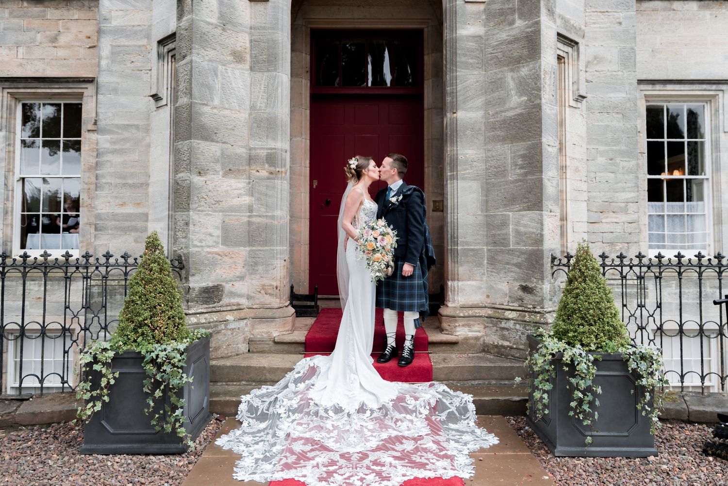 A couple in traditional Scottish wedding attire stands in front of a red door at an elegant stone building entrance.