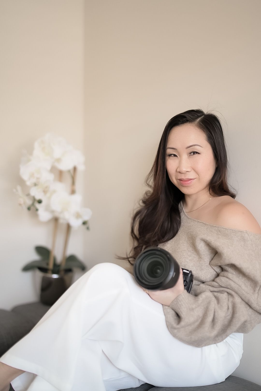 Professional photographer in beige sweater and white pants sits casually with camera lens beside white orchid decor.