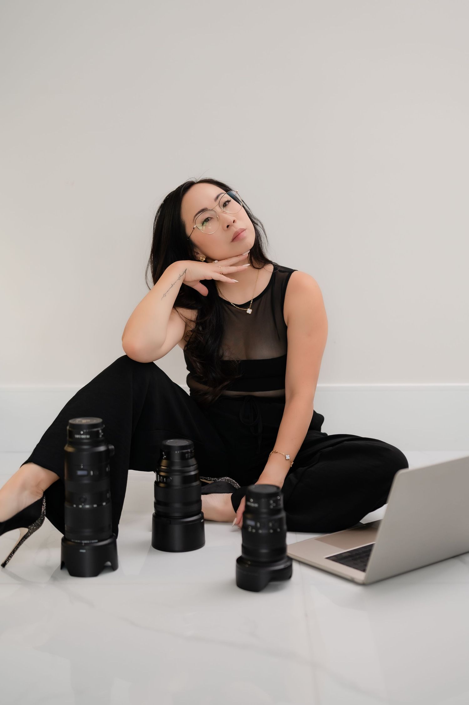Professional photographer in beige sweater and white pants sits casually with camera lens beside white orchid decor.
