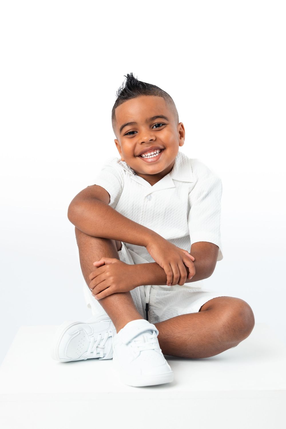Young child wearing white shirt and sneakers poses playfully while lying on a white background.