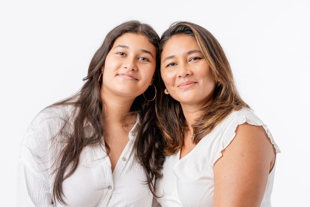 Two people in white tops lean together while smiling against a light background.