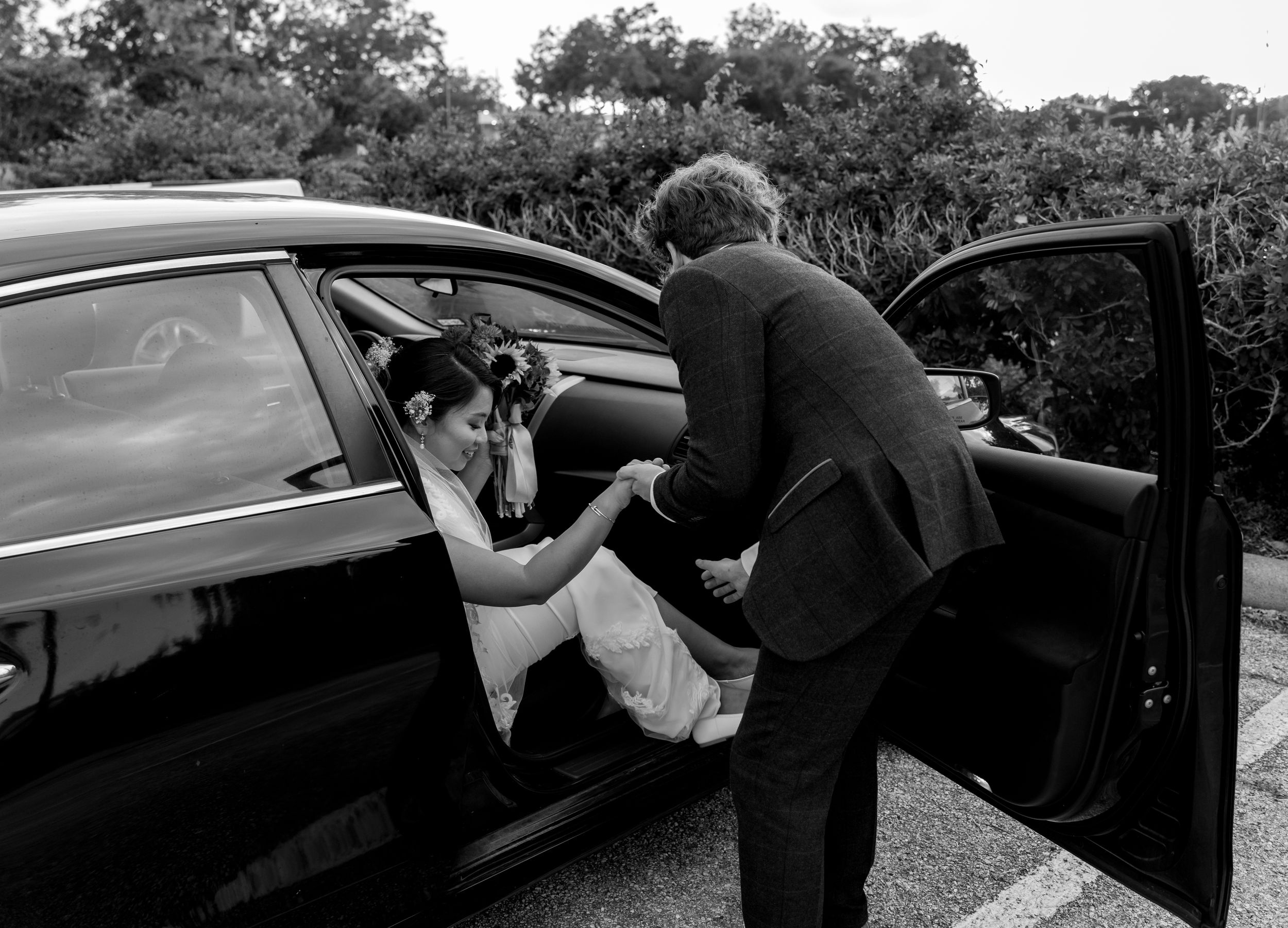 A classic black and white photograph shows someone helping another person exit from a vintage car off-road.