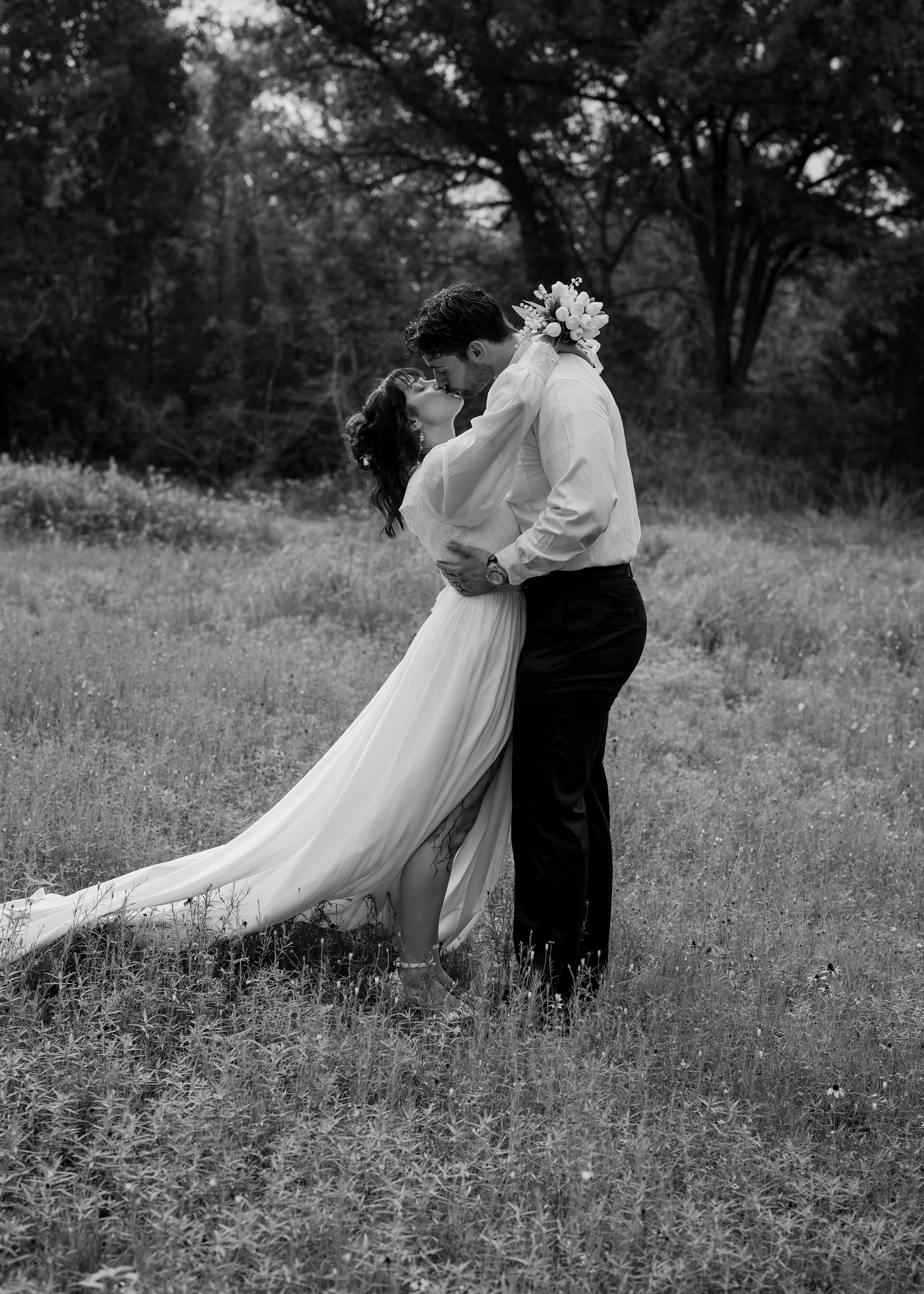 A romantic black and white wedding photo shows a couple embracing in a meadow with flowing dress and trees in background.