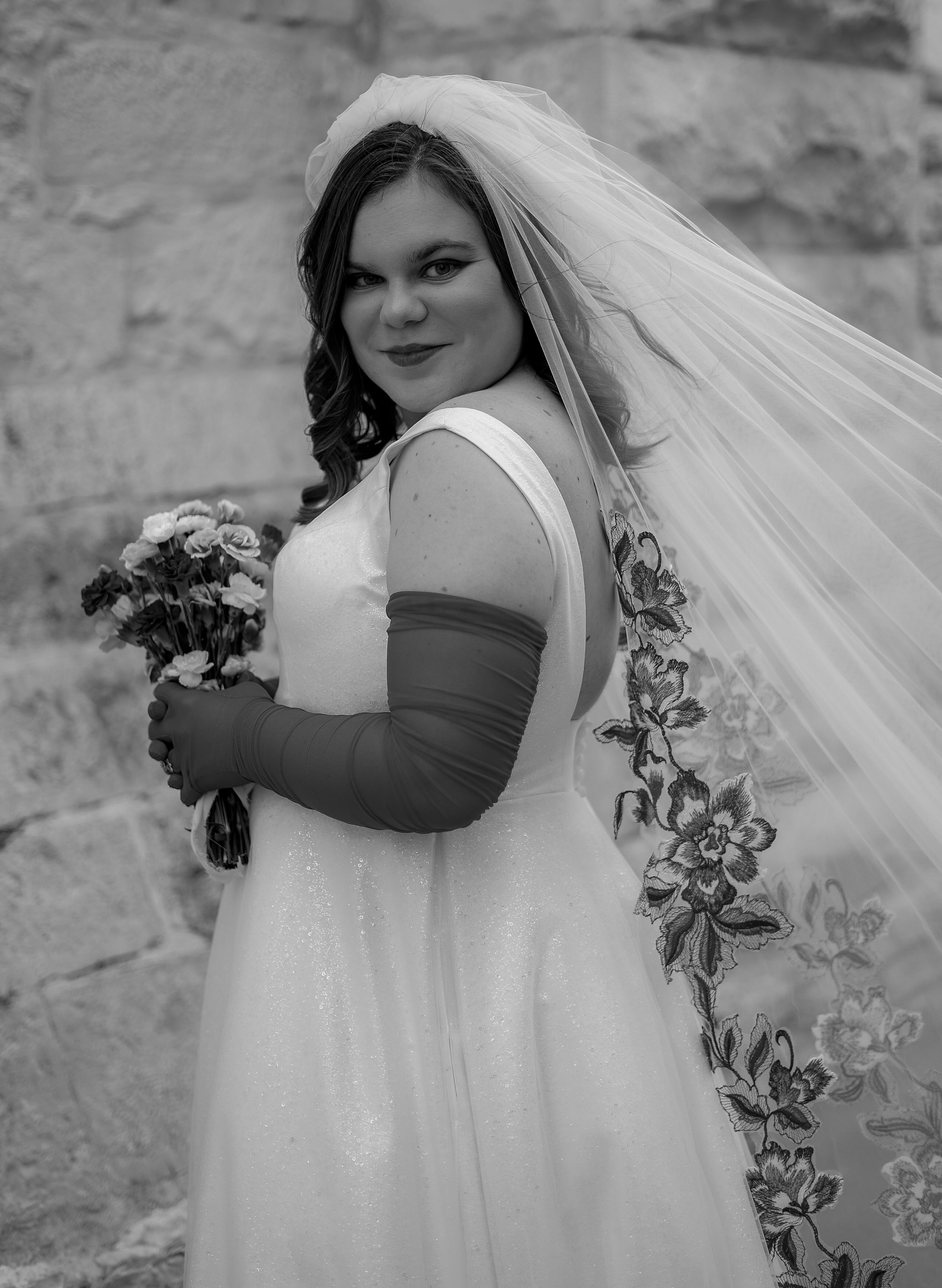 A black and white portrait of a bride in a sleeveless gown holding a bouquet against a stone wall.
