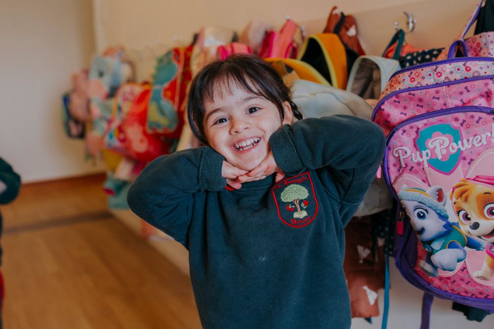 Child smiles while wearing a dark green sweater with backpacks and colorful items visible in the background.