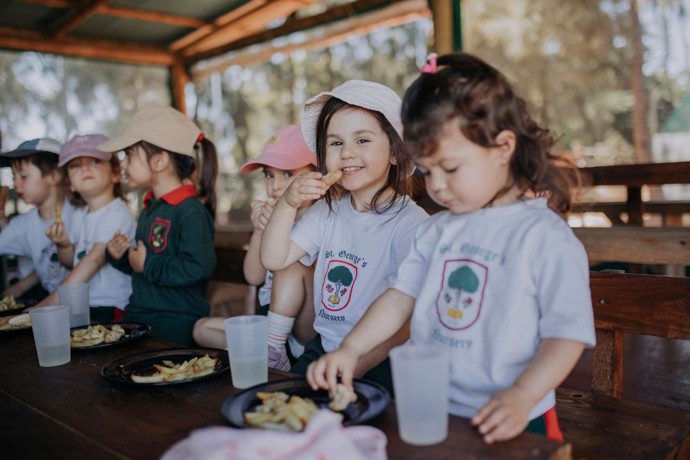 Group of children in matching white t-shirts having a meal together at a covered outdoor table during camp.